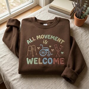May include: A brown sweatshirt featuring the embroidered phrase "ALL MOVEMENT is WELCOME" in pastel hues. The design incorporates images of a walker, wheelchair, and crutches. The sweatshirt is folded on a light-coloured surface.