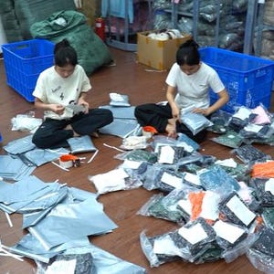 May include: Two individuals in white t-shirts and dark pants sit on a wooden floor, surrounded by numerous plastic-wrapped clothing items and shipping mailers. Blue plastic crates and racks filled with folded garments are visible in the background.