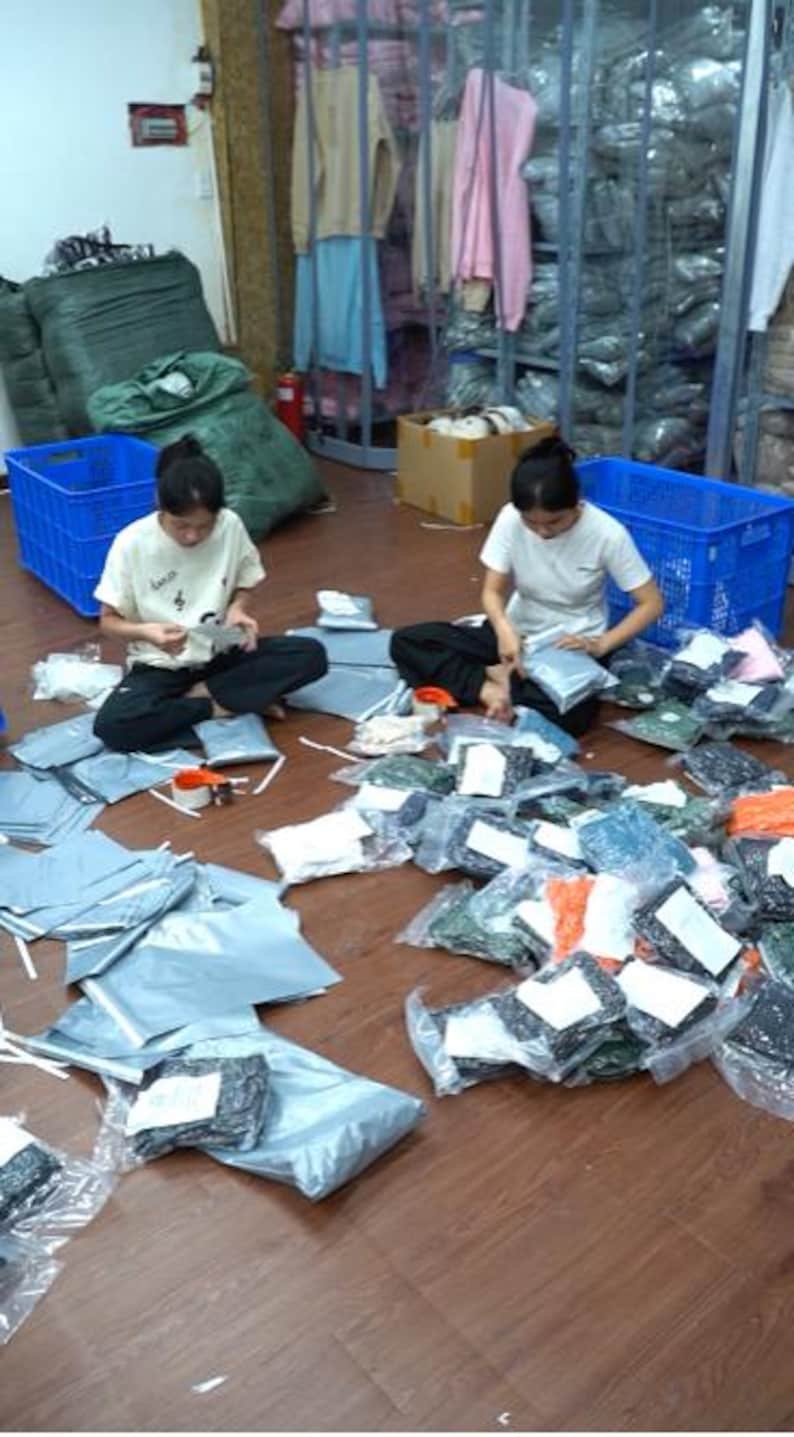 May include: Two individuals in white t-shirts and dark pants sit on a wooden floor, surrounded by numerous plastic-wrapped clothing items and shipping mailers. Blue plastic crates and racks filled with folded garments are visible in the background.