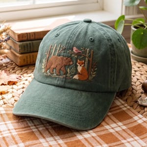 May include: A sage green baseball cap featuring detailed embroidery of a brown bear, a red fox, and a small bird within a forest scene. The cap has a curved brim, stitched eyelets, and a button on top, showcasing a woodland-themed design.