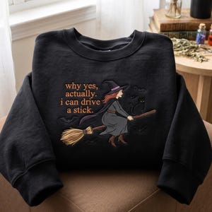 Könnte beinhalten: Ein schwarzes Sweatshirt mit Rundhalsausschnitt und einer Stickerei, die eine Hexe mit rötlichem Haar, spitzem Hut und Umhang auf einem Besenstiel zeigt. Der Text lautet, why yes, actually. i can drive a stick. Kleine Fledermäuse und eine Katze sind abgebildet.