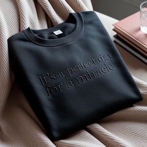 Könnte beinhalten: Dunkelgraues Sweatshirt mit dem gestickten Text "It's a perfect day for a miracle". Das Rundhals-Sweatshirt wird auf einer beigen Decke präsentiert. Bücher und ein Glas Wasser sind im Hintergrund.