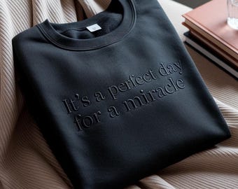 May include: Dark gray sweatshirt with the embroidered text "It's a perfect day for a miracle." The crew neck sweatshirt is displayed on a beige blanket. A stack of books and a glass of water are in the background.
