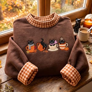 May include: A brown sweatshirt featuring a gingham collar and cuffs, embroidered with four black cats in various mugs. The design includes cats with scarves, a cat spilling a drink, and a cat in a mug next to a small orange pumpkin.
