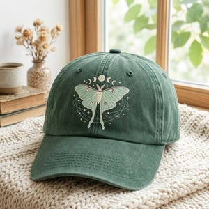 May include: A forest green baseball cap featuring an embroidered light green luna moth centered on the front panel. The moth is surrounded by a circular celestial design including moon phases, thin orbital lines, and small stars in white thread.