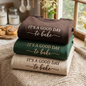 May include: A stack of three crewneck sweatshirts in dark brown, forest green, and beige. Each sweatshirt features the white text "IT'S A GOOD DAY to bake" printed on the front, with the word "bake" in a cursive script font.