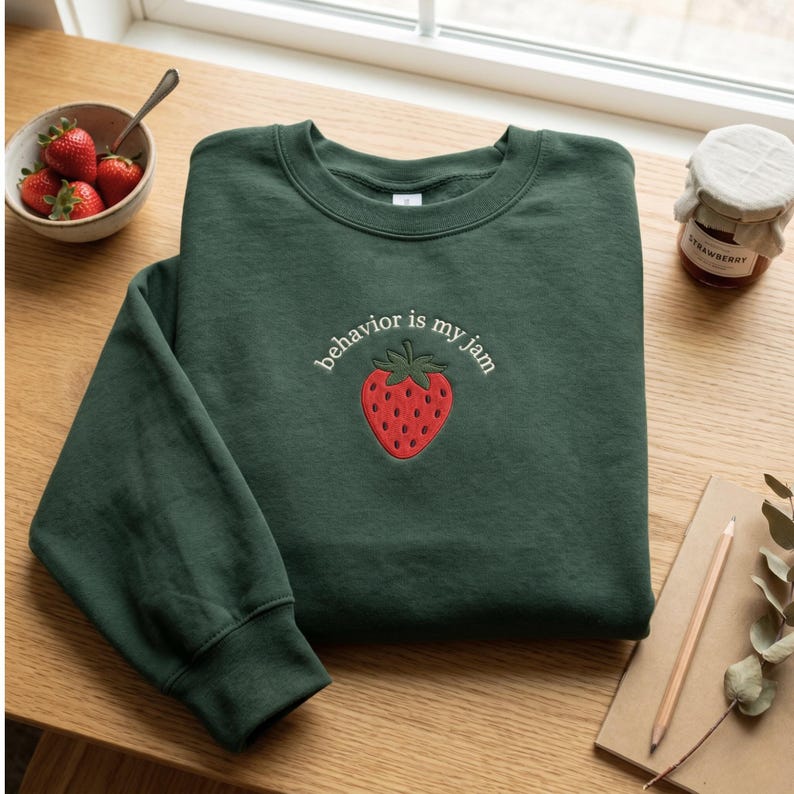 May include: A dark green crewneck sweatshirt featuring an embroidered red strawberry graphic centered on the chest. Above the strawberry, the text "behavior is my jam" is stitched in white thread. The sweatshirt has long sleeves and ribbed cuffs.