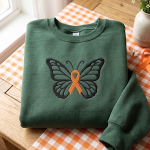 Könnte beinhalten: Ein waldgrüner Rundhals-Pullover mit einer zentralen Stickerei eines schwarz umrandeten Schmetterlings, in dessen Körper ein orangefarbenes Awareness-Band integriert ist. Das Kleidungsstück hat lange Ärmel und einen gerippten Kragen.