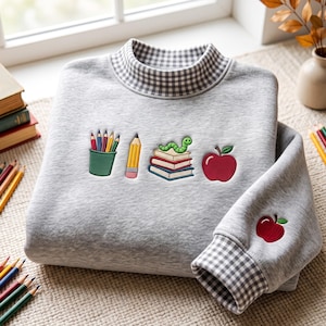 May include: A light grey sweatshirt with a mock neck featuring a grey and white gingham pattern. The front displays embroidered icons of a pencil cup, a yellow pencil, a stack of books with a green worm, and a red apple. A red apple is embroidered on the sleeve.