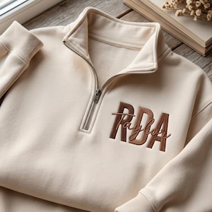 Puede incluir: Una sudadera color crema con cremallera de un cuarto, cuello alto y puños acanalados. En el pecho presenta las letras RDA bordadas en fuente de bloque marrón oscuro, con el nombre Taylor cosido en cursiva marrón sobre las letras.