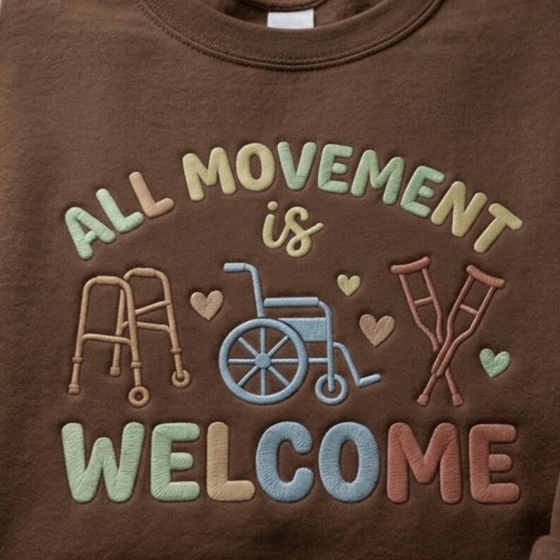 May include: A brown sweatshirt featuring embroidered text and images. The text says "ALL MOVEMENT is WELCOME" in multiple colours. The design includes a walker, wheelchair, crutches, and hearts.