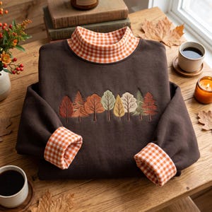 Könnte beinhalten: Ein dunkelbraunes Langarm-Sweatshirt mit Stehkragen und Ärmelbündchen, die mit einem orange-weißen Vichy-Muster gefüttert sind. In der Mitte der Brust befindet sich eine gestickte Reihe verschiedener Bäume in Grün-, Orange- und Gelbtönen.