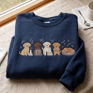 May include: A navy blue crewneck sweatshirt featuring an embroidered design of five puppies in a laboratory setting. The puppies are depicted with scientific equipment including beakers, test tubes, and goggles, with one puppy sleeping in a bed.