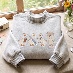 Puede incluir: Una sudadera gris claro con cuello alto y puños con estampado de cuadros vichy en blanco y gris. La parte delantera muestra un bordado de cuatro gansos blancos vestidos como personal médico, sosteniendo un portapapeles, jeringa, estetoscopio y bandeja.