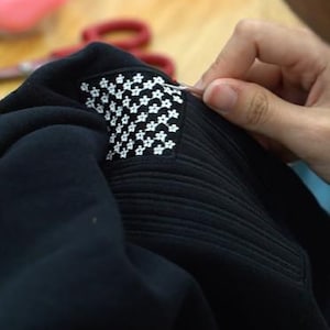 May include: A black fabric garment featuring a rectangular patch with a repeating white floral pattern. The patch is being worked on with a small metal tool, showing detailed stitching and a textured surface on the black material surrounding it.