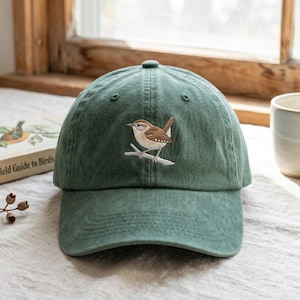 Peut inclure: Une casquette de baseball vert forêt ornée d'une broderie représentant un petit oiseau brun perché sur une branche. La casquette présente une calotte structurée, une visière incurvée et des œillets cousus sur les panneaux supérieurs.