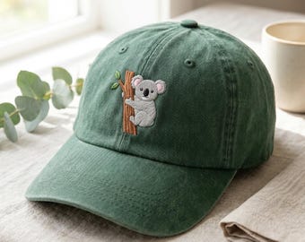 Puede incluir: Una gorra de béisbol de color verde oscuro con un diseño bordado de un koala gris claro aferrado a un tronco de árbol marrón con una hoja verde. La gorra tiene una corona estructurada, una visera curva y ojales cosidos para ventilación.
