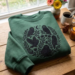 May include: A forest green crewneck sweatshirt featuring a dark black embroidered design of four owls perched on branches and flying amidst floral elements, leaves, and a crescent moon with stars centered on the front of the garment.