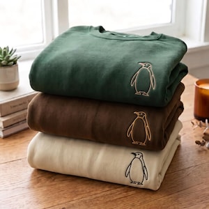 Könnte beinhalten: Ein Stapel aus drei Rundhals-Sweatshirts in Waldgrün, Dunkelbraun und Creme. Jedes Sweatshirt ist mit einer einfachen, minimalistischen Stickerei eines Pinguin-Umrisses im oberen linken Brustbereich versehen. Die Kleidungsstücke sind ordentlich gestapelt.