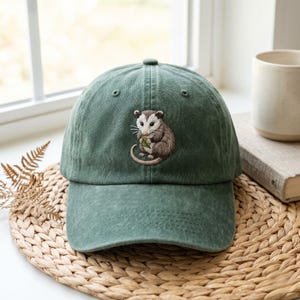 Op de afbeelding: Een bosgroene baseballpet met een geborduurde afbeelding van een kleine opossum die een groen blaadje vasthoudt. De pet heeft een gestructureerde kroon, een gebogen klep en een klassiek ontwerp met zes panelen en gestikte oogjes bovenop.