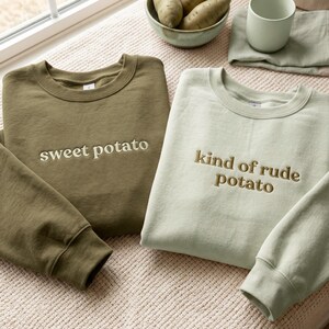 Op de afbeelding: Twee sweaters met ronde hals in olijfgroen en saliegroen. De olijfgroene sweater heeft de geborduurde tekst "sweet potato" in een crèmekleur. De saliegroene sweater heeft de geborduurde tekst "kind of rude potato" in bruin.