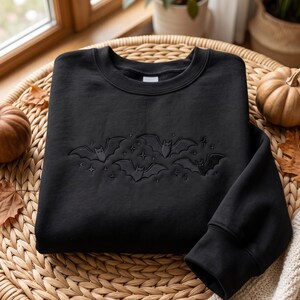 Peut inclure: Un sweat-shirt noir à col rond orné d'une broderie centrale représentant trois chauves-souris en vol entourées de petites étoiles à quatre branches. La broderie est réalisée avec un fil noir assorti, créant un effet ton sur ton.