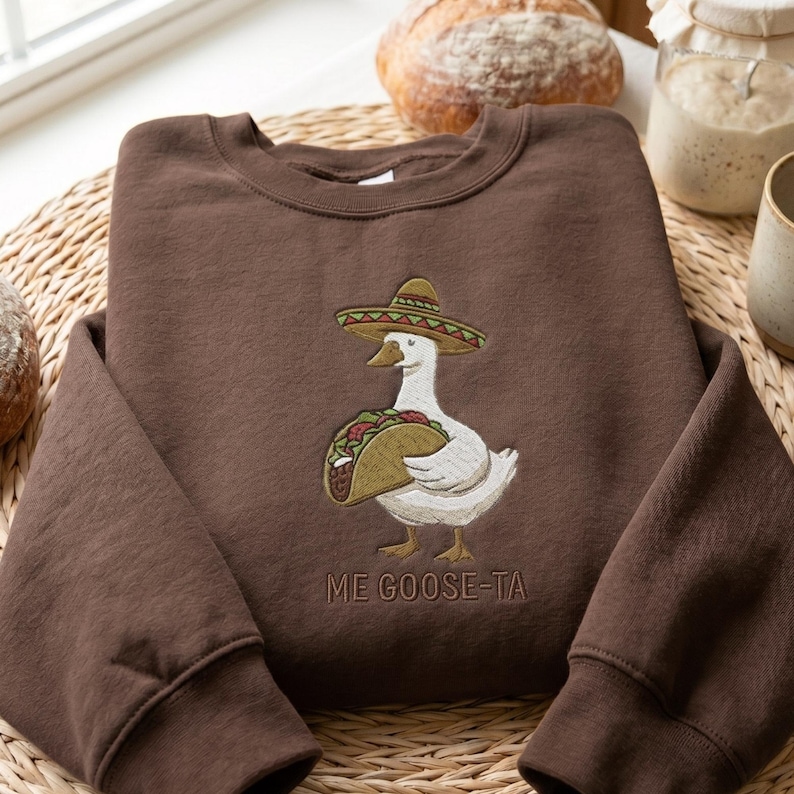 May include: A brown sweatshirt featuring an embroidered design of a white goose wearing a sombrero and holding a taco. The text "ME GOOSE-TA" is embroidered below the goose. The sweatshirt is made of a soft material.