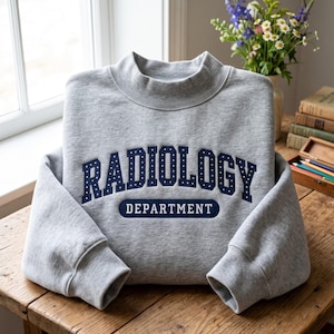 Puede incluir: Una sudadera gris claro de cuello alto con letras bordadas en azul oscuro que dicen RADIOLOGY DEPARTMENT. La palabra RADIOLOGY está arqueada y decorada con pequeños puntos blancos, mientras que la palabra DEPARTMENT está en un óvalo azul oscuro.