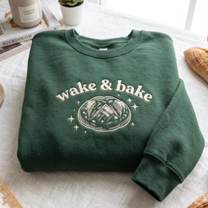 May include: A forest green crewneck sweatshirt featuring an embroidered design on the chest. The embroidery includes the text "wake & bake" in cream-colored thread above a detailed illustration of a round loaf of sourdough bread with star accents.