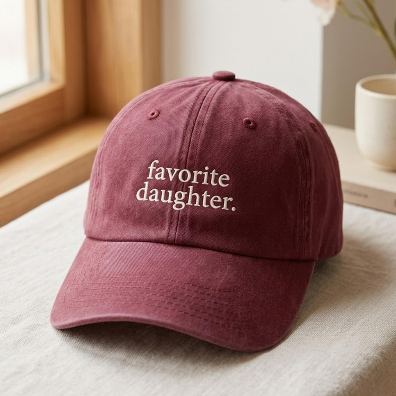 May include: A maroon baseball cap featuring the embroidered text favorite daughter. in white lowercase letters. The cap has a structured crown, a curved brim, and a button at the top center, showcasing a classic six-panel design with stitching.