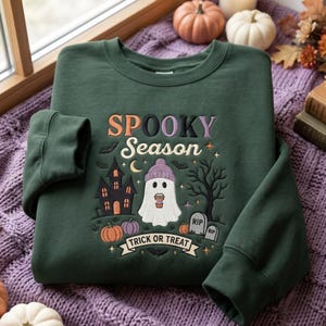 Peut inclure: Un sweat-shirt à col rond vert foncé avec une broderie représentant le texte "SPOOKY Season" et "TRICK OR TREAT". Le motif comprend un fantôme portant un bonnet violet, une maison hantée, un arbre dénudé, des citrouilles et des pierres tombales.
