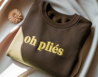 May include: A dark brown crewneck sweatshirt featuring the text "oh pliés" embroidered in yellow thread across the chest. The garment has a ribbed collar and long sleeves, showcasing a clean, simple design with bold, contrasting lettering.