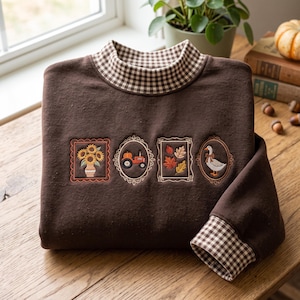 Puede incluir: Una sudadera de manga larga de color marrón oscuro con cuello alto y puños con un patrón de cuadros vichy en marrón y blanco. El frente muestra cuatro marcos bordados, rectangulares y ovalados, con un jarrón de girasoles, un tractor, hojas de otoño y un ganso.