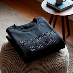 Op de afbeelding: Zwart sweatshirt met de geborduurde tekst "It's a good day to do math". Het sweatshirt ligt opgevouwen op een geweven poef. Een kleine houten bijzettafel met een boek en een mok staat op de achtergrond.
