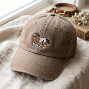 Puede incluir: Una gorra de béisbol marrón con un diseño bordado de un caballo corriendo, con una mancha blanca en la cara y manchas blancas en los cuartos traseros. La gorra tiene una visera curva, un botón en la parte superior y ojales cosidos.