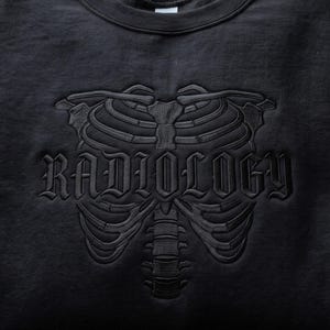 May include: A black crewneck sweatshirt featuring a tonal black embroidered design of a human rib cage and spine. The word RADIOLOGY is embroidered in a gothic-style font across the center of the rib cage, matching the black fabric.
