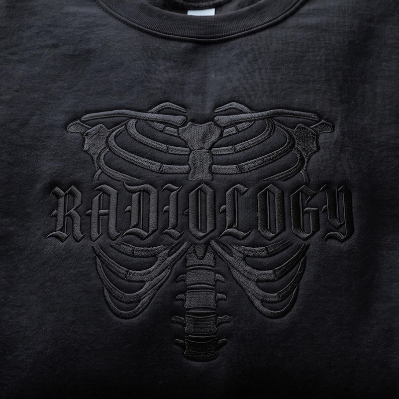 May include: A black crewneck sweatshirt featuring a tonal black embroidered design of a human rib cage and spine. The word RADIOLOGY is embroidered in a gothic-style font across the center of the rib cage, matching the black fabric.