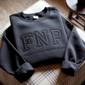 Könnte beinhalten: Ein dunkelgraues Sweatshirt mit den gestickten Buchstaben "FNP" im College-Stil. Unter den Buchstaben sind die Worte "FAMILY NURSE PRACTITIONER" gestickt. Das Sweatshirt ist ordentlich auf einer gemusterten Oberfläche gefaltet.