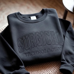 Puede incluir: Una sudadera gris oscuro con las palabras "SURGICAL TECHNOLOGIST" bordadas en un tono a juego. La sudadera está doblada sobre una superficie texturizada, con las mangas caídas casualmente.
