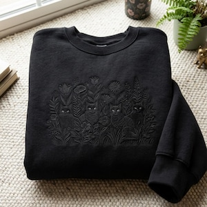 May include: A black crewneck sweatshirt featuring tonal black embroidery on the chest. The design depicts four cats nestled among various flowers, including tulips and daisies, creating a textured, monochromatic botanical scene.