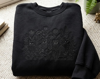 May include: A black crewneck sweatshirt featuring tonal black embroidery on the chest. The design depicts four cats nestled among various flowers, including tulips and daisies, creating a textured, monochromatic botanical scene.