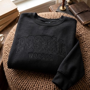 Könnte beinhalten: Ein schwarzes Sweatshirt mit Rundhalsausschnitt und dem in passendem schwarzem Garn gestickten Schriftzug POSTAL WORKER. Das Wort POSTAL ist in einer großen College-Schriftart gehalten, in die zarte florale Linienzeichnungen integriert sind.