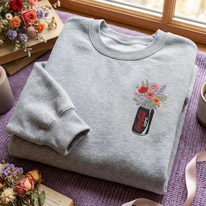 Könnte beinhalten: Ein graumeliertes Sweatshirt mit Rundhalsausschnitt und einer Stickerei auf der linken Brust. Die Stickerei zeigt eine schwarze Getränkedose mit der Aufschrift Coca-Cola Zero und einem bunten Blumenstrauß aus roten, orangen, lila und weißen Blüten.