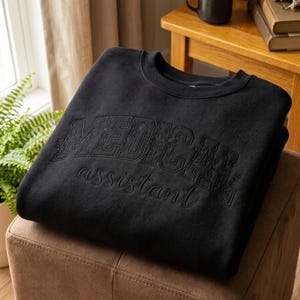 以下が含まれることがあります： 黒いクルーネックのスウェットシャツ。胸元には花柄のディテールが施された大きなブロック体の刺繍で「MEDICAL」とあり、そのすぐ下に筆記体の刺繍で「assistant」という文字がデザインされています。