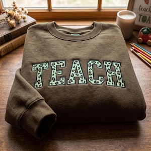 Könnte beinhalten: Ein dunkelbraunes Sweatshirt mit Rundhalsausschnitt und dem Wort TEACH in großen Blockbuchstaben auf der Brust. Die Buchstaben sind hellgrün mit einem dunkelbraunen Leopardenmuster und mit dunkelbraunem Garn umrandet.