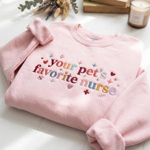 May include: A light pink crewneck sweatshirt featuring multi-colored embroidered text that reads your pet's favorite nurse. The design includes small embroidered hearts, stars, and medical-themed symbols scattered around the lettering.
