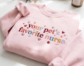May include: A light pink crewneck sweatshirt featuring multi-colored embroidered text that reads your pet's favorite nurse. The design includes small embroidered hearts, stars, and medical-themed symbols scattered around the lettering.
