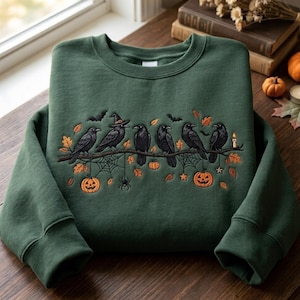 May include: A dark forest green crewneck sweatshirt featuring an embroidered design of six black crows perched on a branch. The scene includes orange pumpkins, a spider, spiderwebs, bats, autumn leaves, and a small lit candle in warm tones.