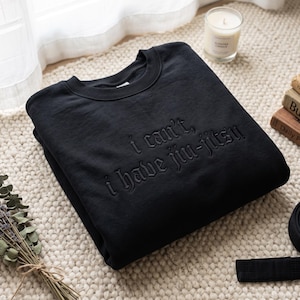 Puede incluir: Una sudadera negra de cuello redondo con el texto bordado I can't, I have jiu-jitsu en una fuente gótica. La prenda tiene mangas largas, un cuello acanalado y un color negro sólido en toda la tela, mostrando un diseño limpio y sencillo.