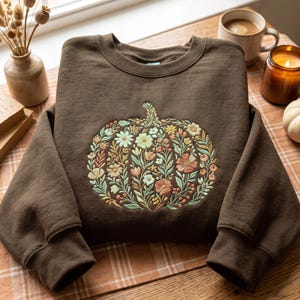 May include: A dark brown crewneck sweatshirt featuring a large, central embroidered design of a pumpkin composed entirely of various small flowers, leaves, and stems in shades of sage green, muted orange, cream, and soft yellow.